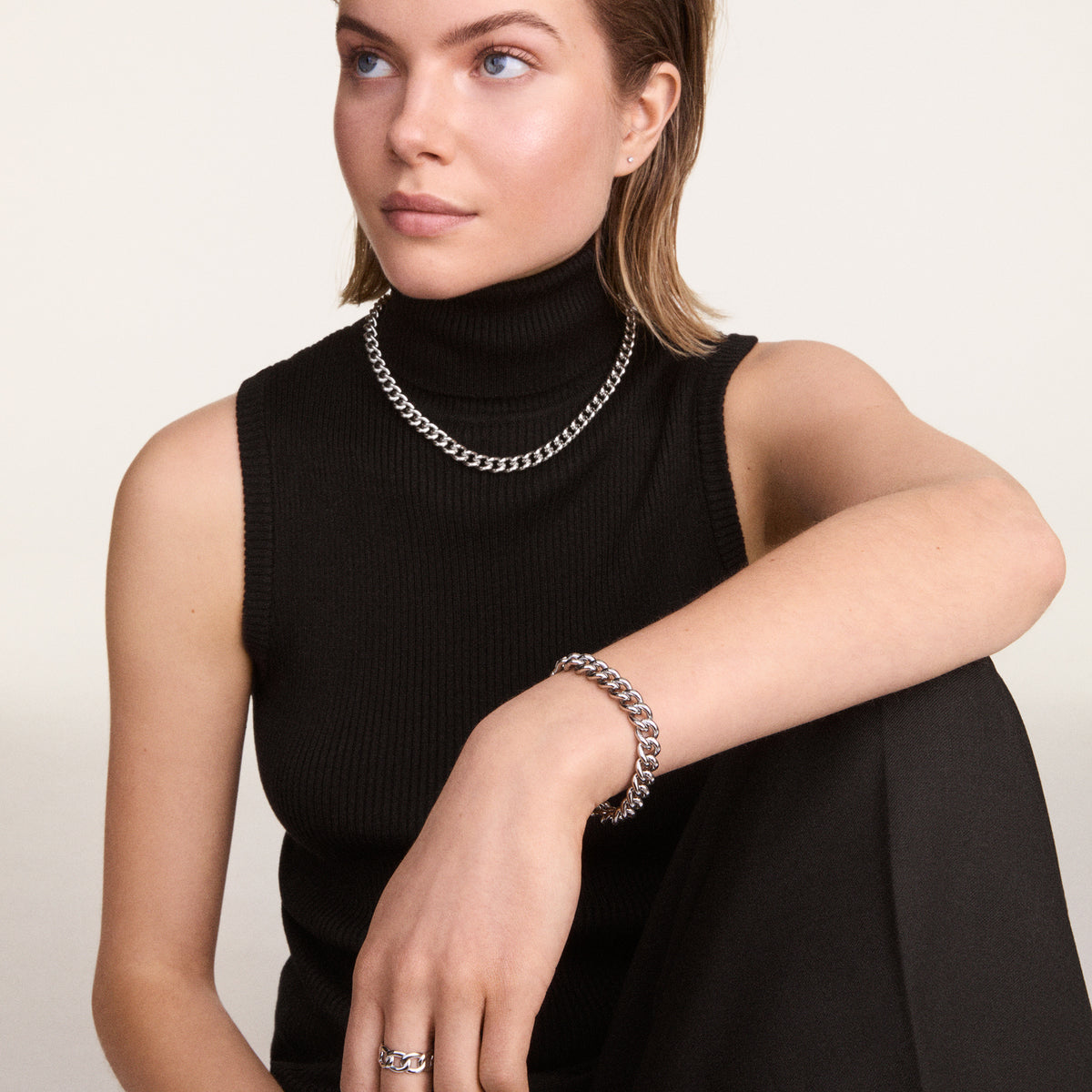 Person wearing a chunky silver chain ring on their finger, styled with a matching chain bracelet and necklace, paired with a sleeveless black turtleneck top for a modern, coordinated look.