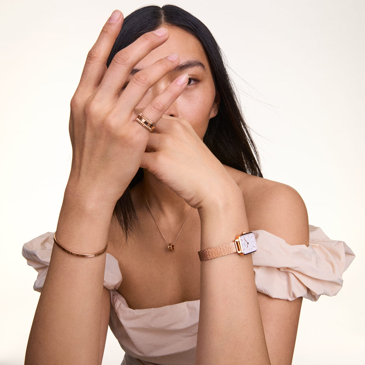A woman wearing the Elevation Ring Rose Gold on her finger, styled with a matching rose gold bracelet, necklace, and watch, paired with an off-shoulder cream top.