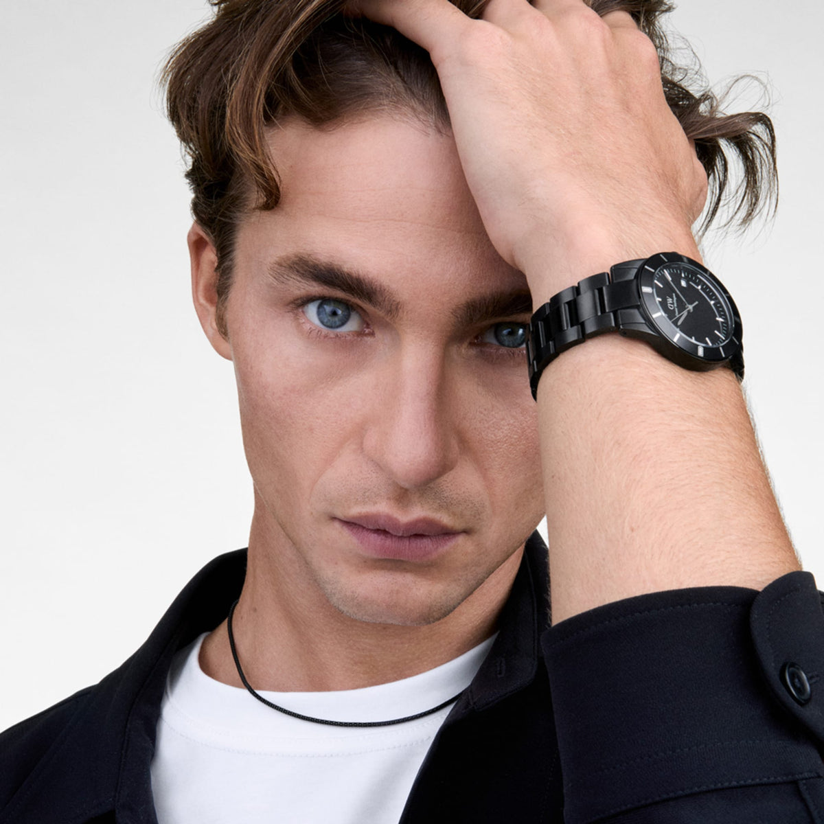 A man wearing the Daniel Wellington Iconic Paradigma Link Black watch, showcasing its sleek black dial, satin matte bezel, and polished 3-link stainless steel bracelet styled with a white T-shirt and dark shirt.