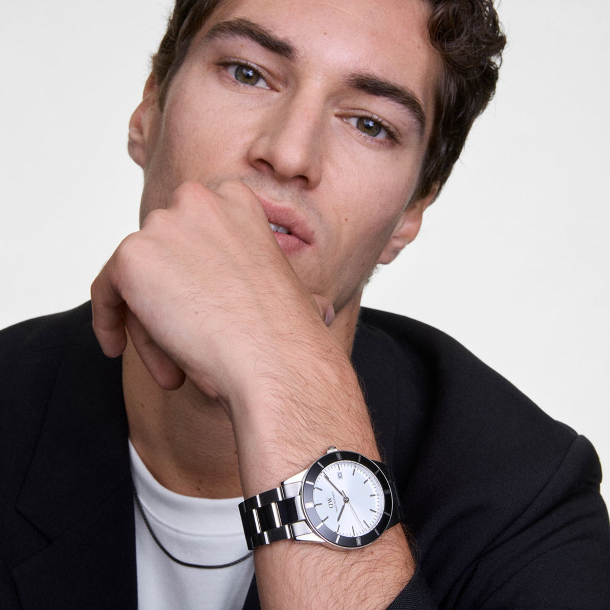 Man wearing the Daniel Wellington Iconic Paradigma silver watch with a black bezel and stainless steel bracelet, styled with a black mesh necklace and a tailored black jacket for a refined, modern look.
