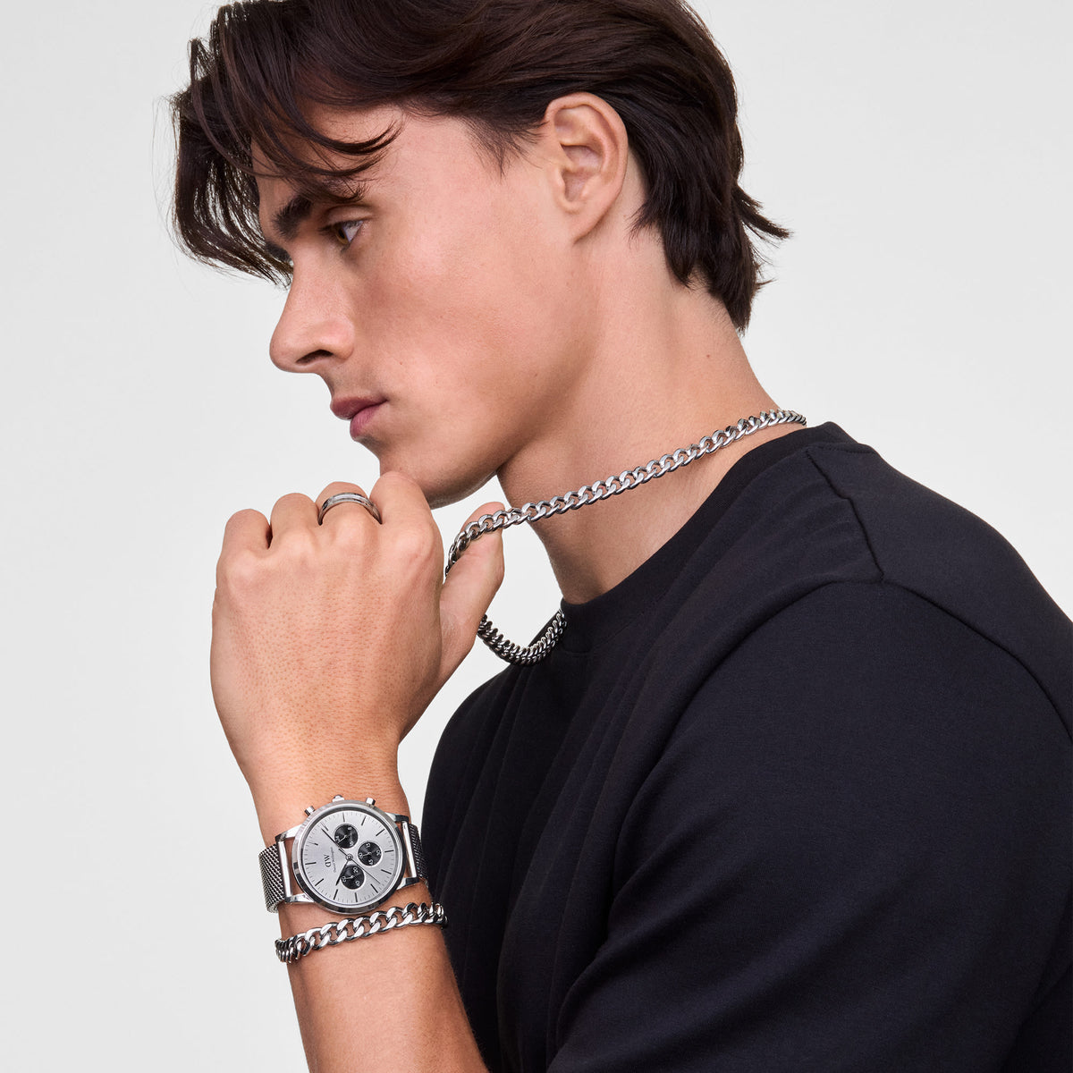 A person wearing the Ellipse Cuban Chain Bracelet Silver on their wrist, styled with a matching silver watch and chain necklace, showcasing the bracelet’s bold, polished links and contemporary design against a black top.