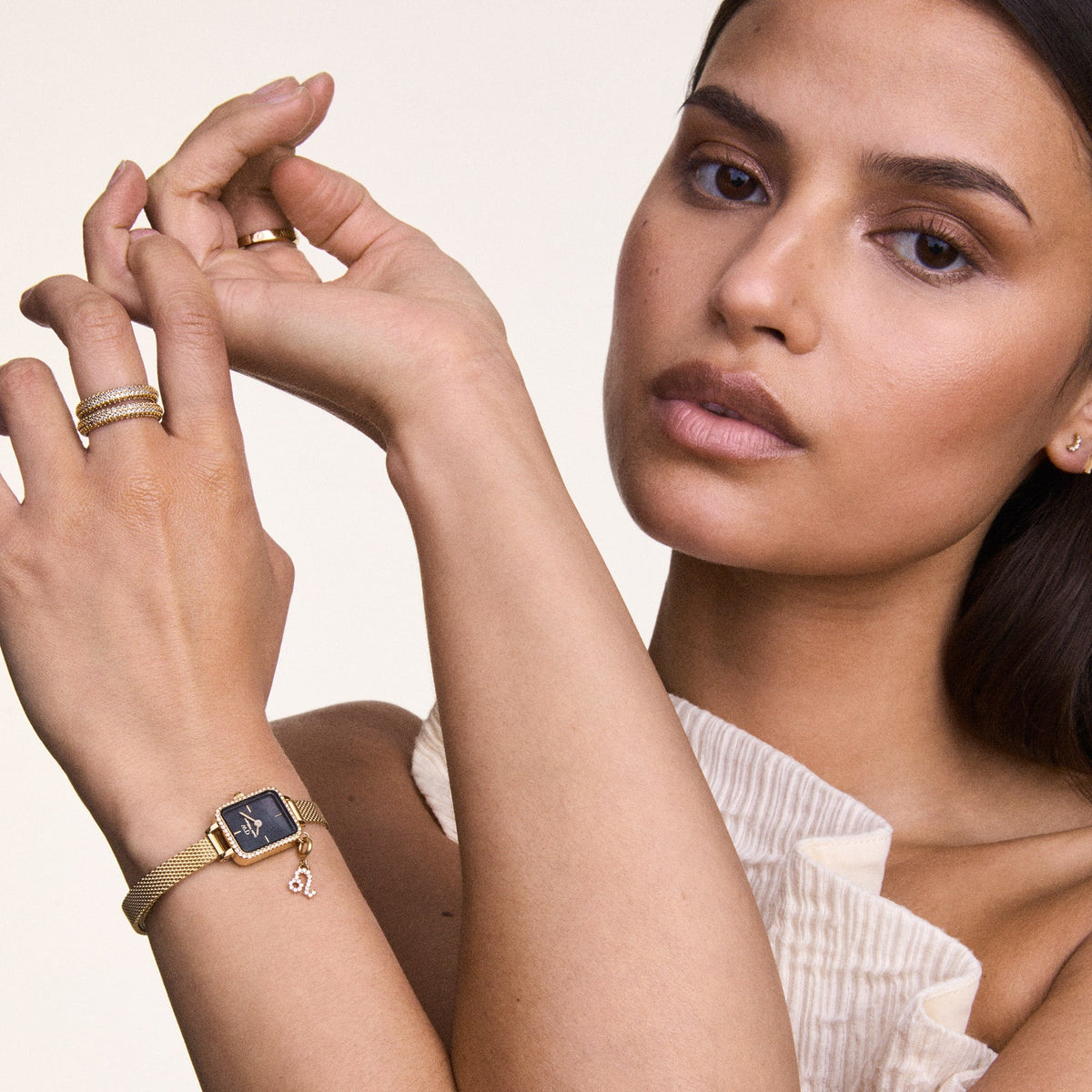 A woman wearing the Quadro Mini Crystal Zodiac Evergold watch, featuring a gold mesh strap, rectangular crystal-set case, black mother of pearl dial, and a sparkling zodiac charm, styled with gold rings for an elegant and refined look.
