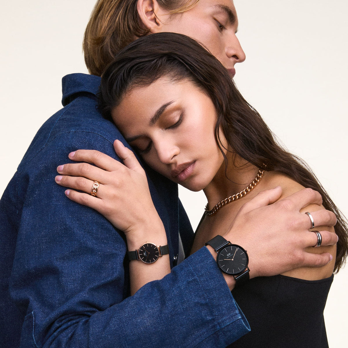 A woman wearing the Petite Ashfield watch with a matte black dial and rose gold case, styled with a black mesh strap, embracing a man also wearing a black mesh strap watch.