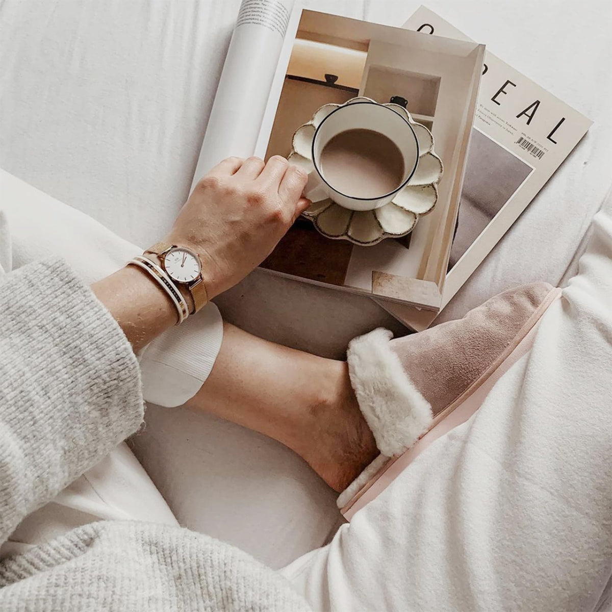 A person relaxing in cosy loungewear wears the Emalie Bracelet Satin White Rose Gold alongside a Daniel Wellington watch, holding a cup of coffee while reading a magazine.