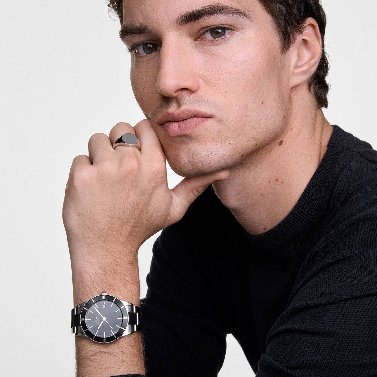 Man wearing the Daniel Wellington Iconic Paradigma Black watch with a silver link strap and a classic silver signet ring, styled with a black jumper for a modern, sophisticated look.