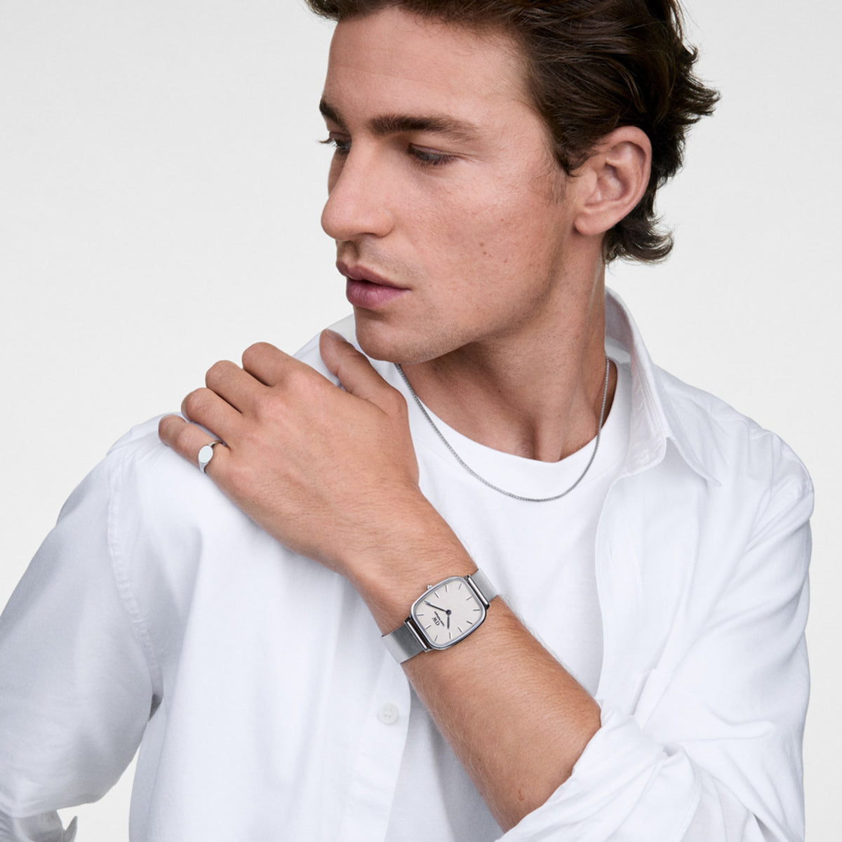 A person wearing the Daniel Wellington Marlon Wire Linen Silver watch, styled with a crisp white shirt and silver jewellery, highlighting the cushion-shaped case, linen-textured dial, and sleek silver mesh strap.