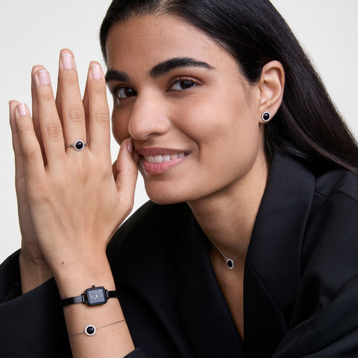 A woman wearing the Quadro Mini Reflection Black watch with a polished black rectangular case and mesh strap, styled alongside matching black jewellery including a ring, bracelet, necklace, and earrings, all set against a black blazer for a sophisticated, coordinated look.