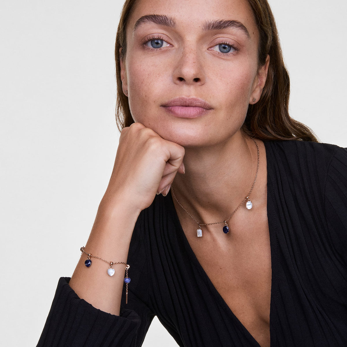 Woman wearing a rose gold necklace and bracelet, each adorned with the Blue Spot Oval Rose Gold Charm and other semi-precious stone charms, styled with a black V-neck top for an elegant, layered jewellery look.