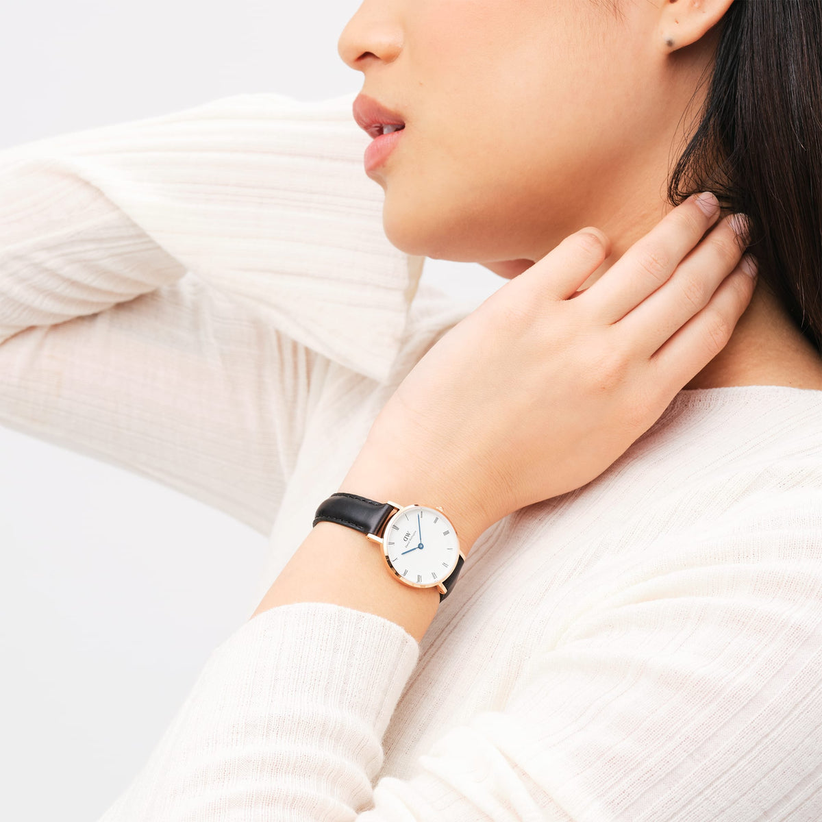 A woman wearing the Petite Roman Numerals Sheffield Rose Gold watch, featuring a white dial with Roman numerals, blue hands, a rose gold case, and a black leather strap, styled with a light textured top.