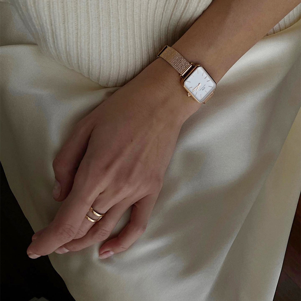 A hand wearing the Elan Dual Ring Rose Gold with a matching rose gold mesh strap watch, styled against a cream satin skirt and ribbed knit top.