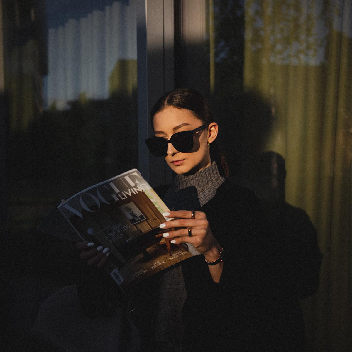 A person wearing Grande Bio-Acetate Black sunglasses reads a magazine outdoors, styled with a dark coat and grey roll-neck for a sophisticated, modern look.