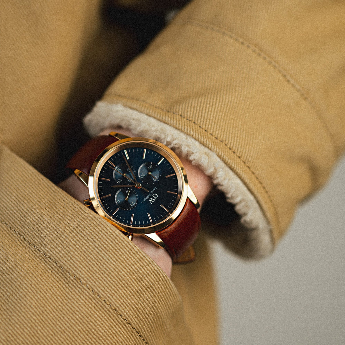 A man wears the Daniel Wellington Iconic Chronograph St Mawes Arctic Rg watch, featuring a rose gold case, blue sunray dial with three sub-dials, and a brown leather strap, styled with a tan shearling-lined coat.