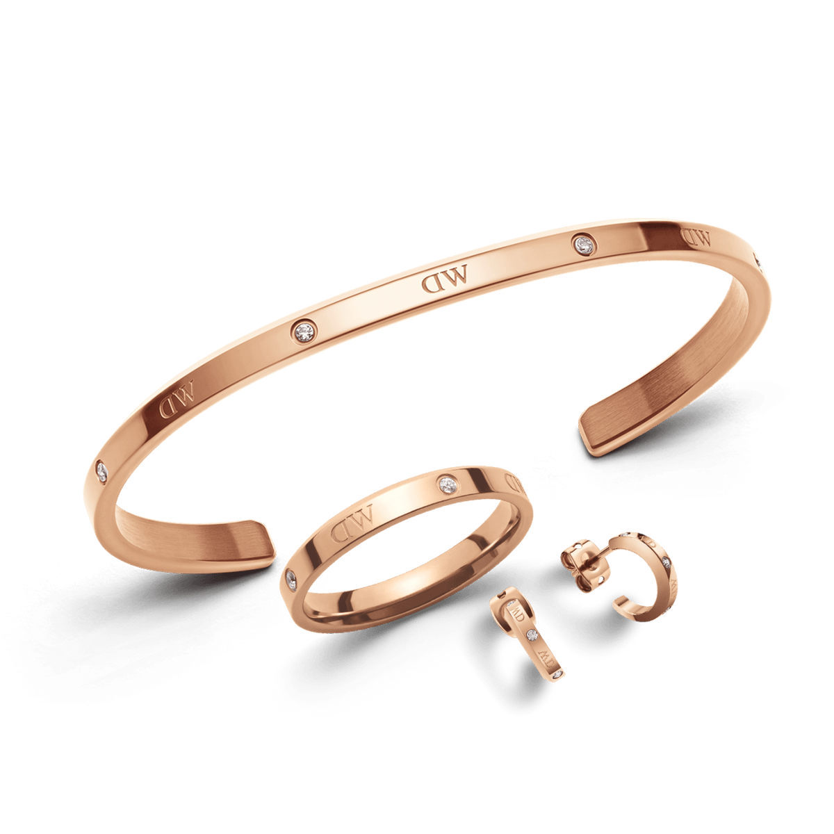 Rose gold jewellery set featuring a sleek open bracelet, matching ring, and small hoop earrings, each adorned with subtle Daniel Wellington branding and delicate crystal accents. The polished finish and coordinated design create an elegant, modern look.