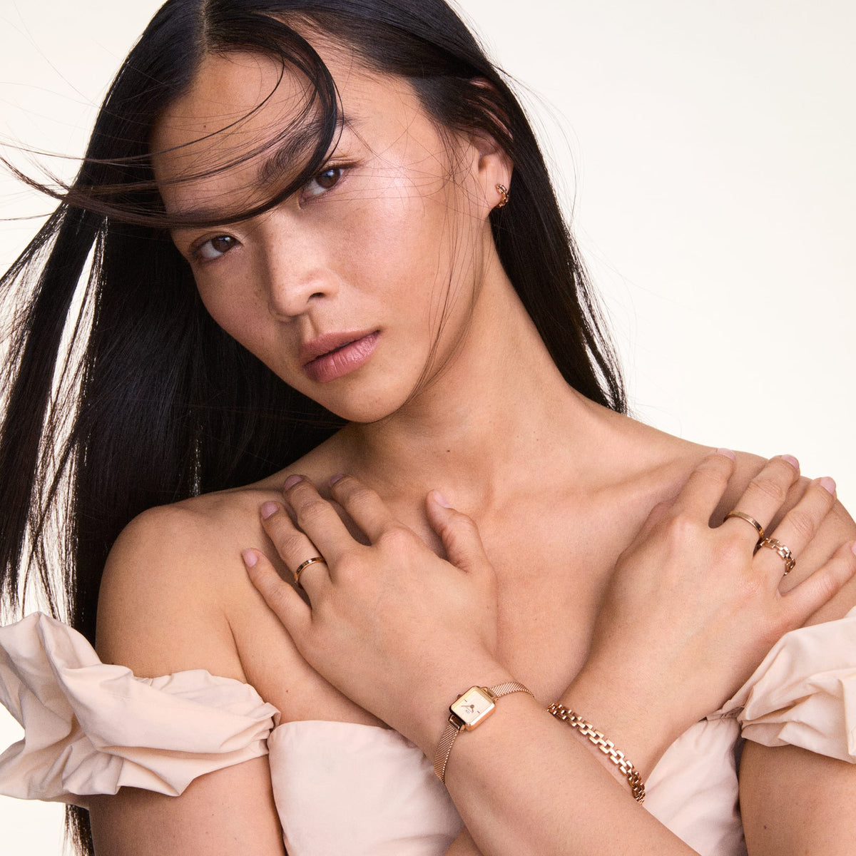 A woman wearing the Quadro Mini Melrose Champagne watch with a petite rectangular champagne dial and rose gold mesh strap, styled with matching rose gold jewellery and an off-shoulder cream dress.