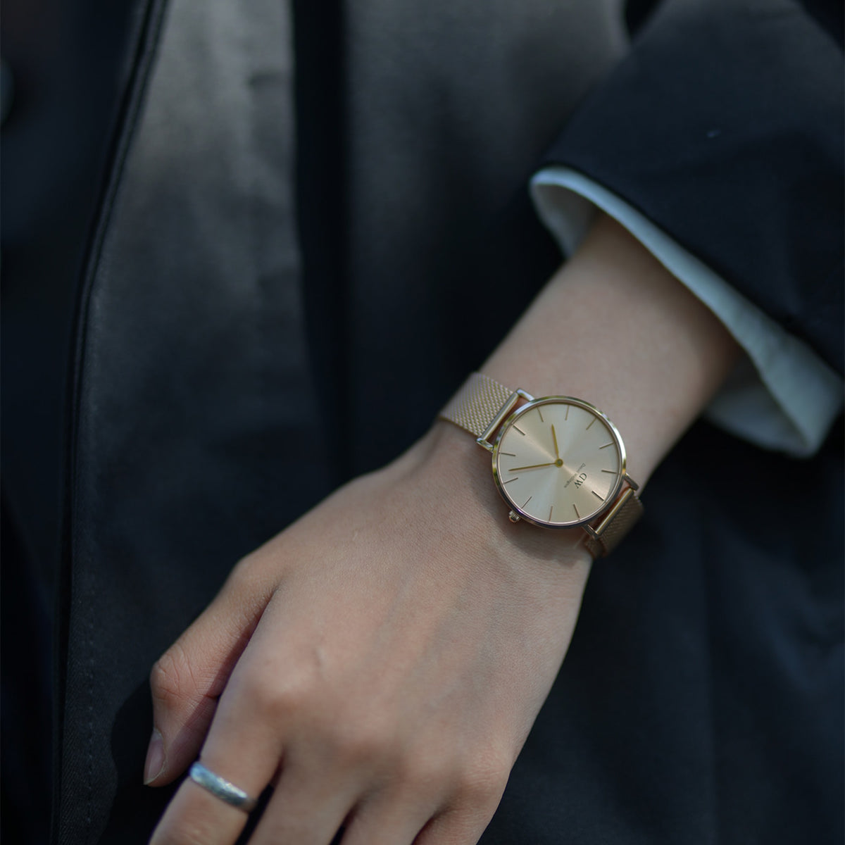 Petite Unitone Gold women’s watch with gold mesh strap and minimalist dial, styled on wrist with a dark jacket.