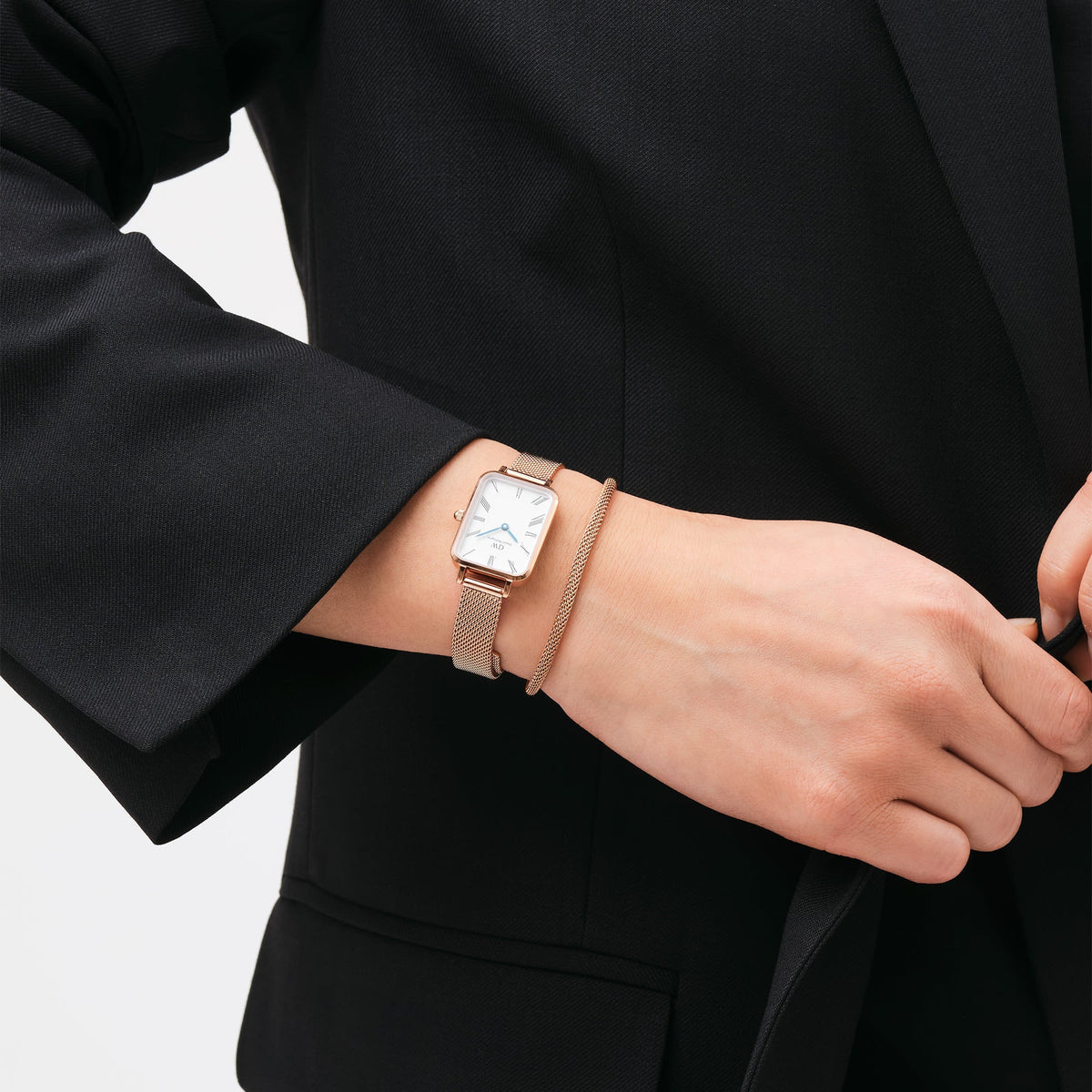 A woman wearing the Quadro Roman Numerals Melrose rose gold rectangular watch with a white dial, Roman numeral markers, and a matching mesh bracelet, styled with a black blazer for a sophisticated look.