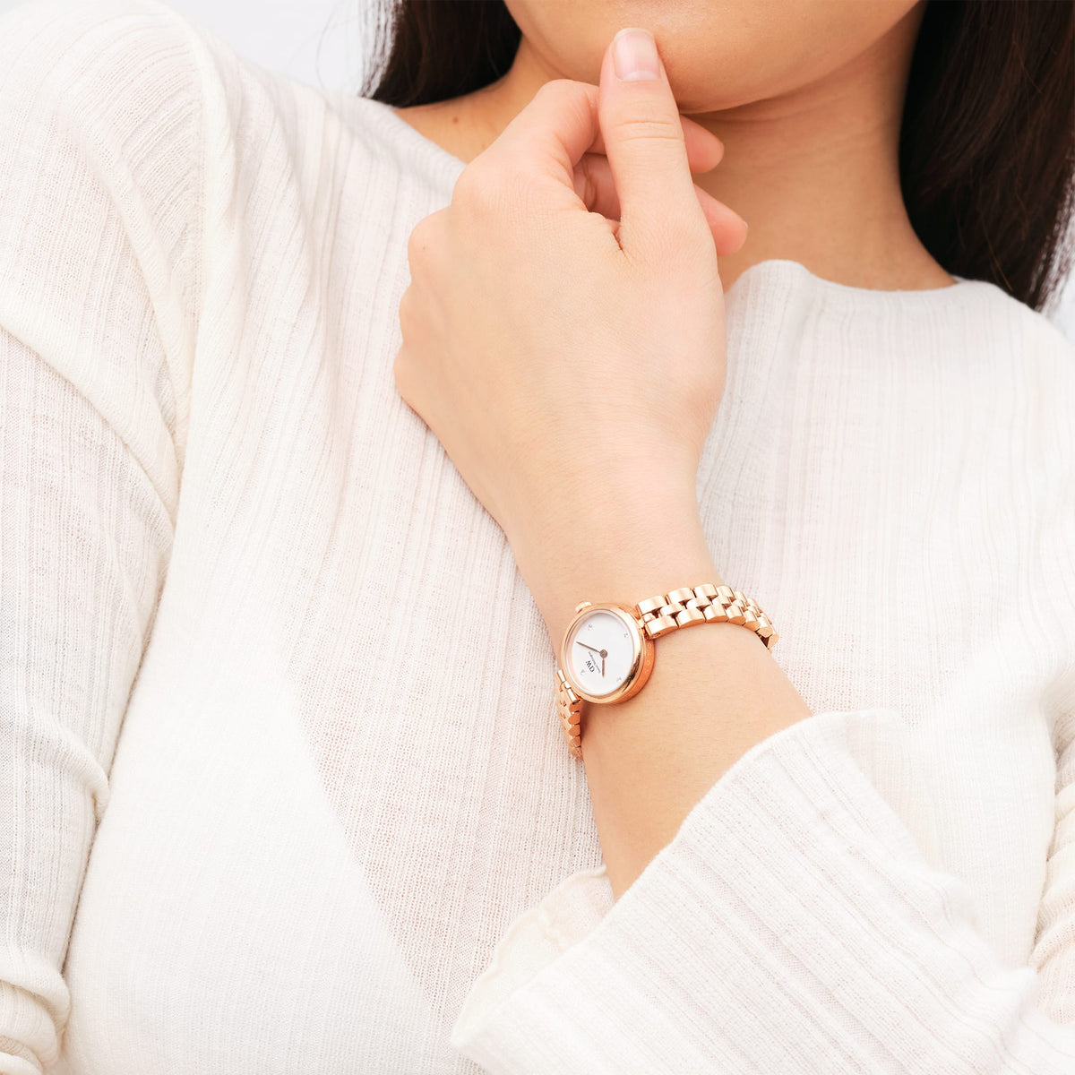 A woman wearing the Elan Lumine Rose Gold watch, featuring a polished rose gold three-link bracelet and a minimalist white dial with crystal markers, styled with a light textured blouse.