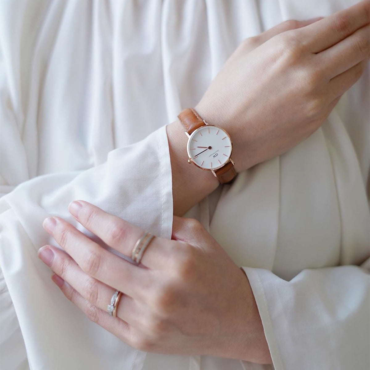 A woman wears the Daniel Wellington Petite Durham Rose Gold watch with a light brown leather strap and minimalist white dial, styled with delicate rings and a white blouse.