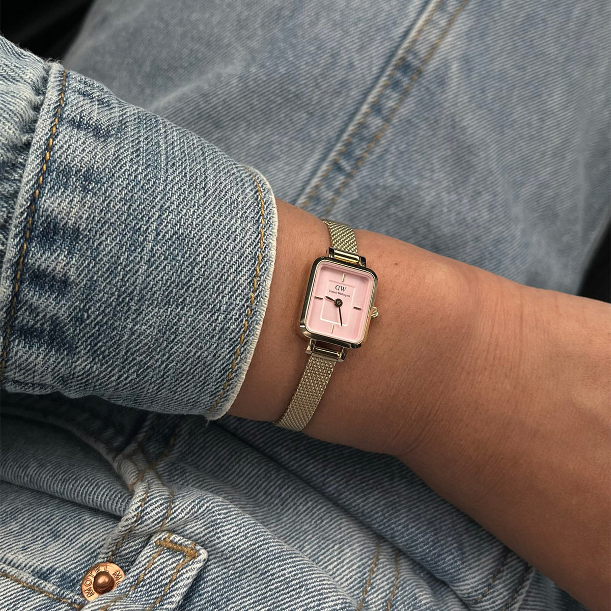 Quadro Mini Evergold Blush women’s watch with a rectangular gold case, blush pink dial, and gold mesh strap, styled on a wrist with casual denim clothing.