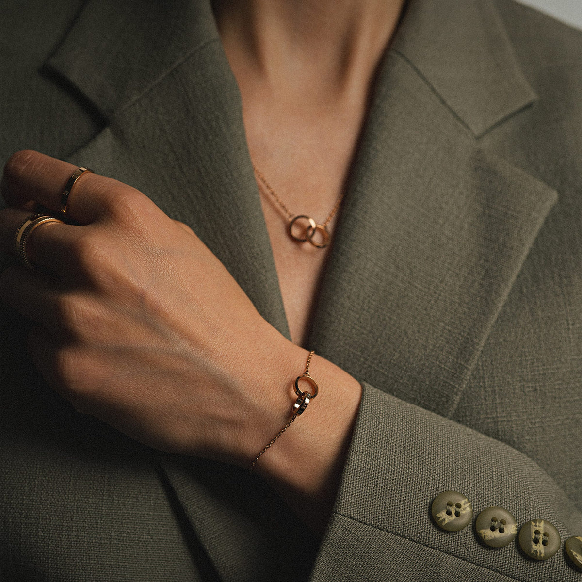 A person wearing the rose gold Elan Unity Bracelet with two interlocking rings on a fine chain, styled with a matching necklace and gold rings for a sophisticated, coordinated look.