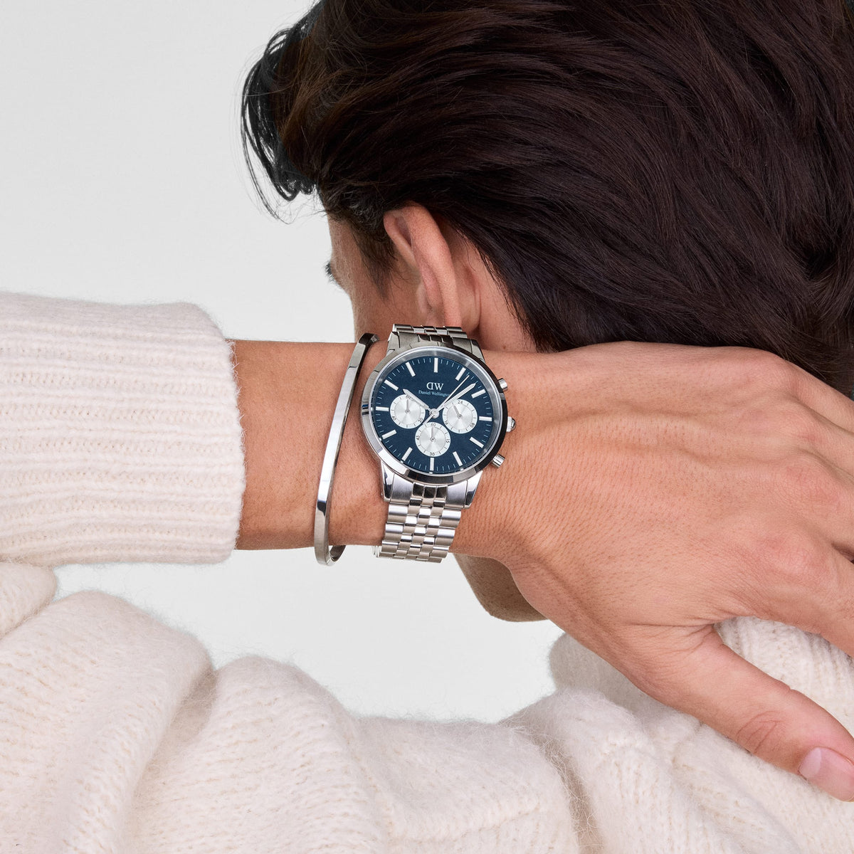 A person wearing the Daniel Wellington Classic Bangle Silver alongside a stainless steel watch, highlighting the bracelet’s polished finish and minimalist design against a cream knit jumper.