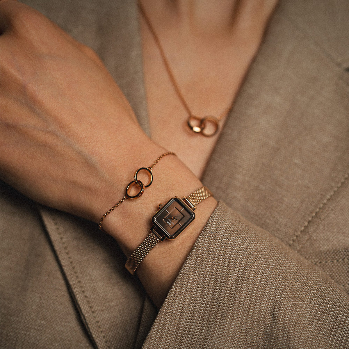 Woman wearing the Quadro Mini Melrose Amber watch with a rectangular amber dial and rose gold mesh strap, styled alongside matching rose gold jewellery and a neutral blazer for an elegant, coordinated look.