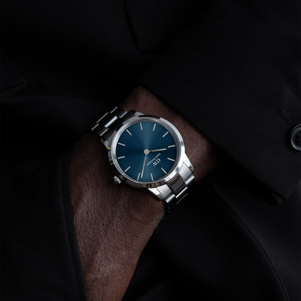 The Daniel Wellington Iconic Link Arctic watch with a blue sunray dial and silver stainless steel bracelet, styled on the wrist with a black suit jacket.