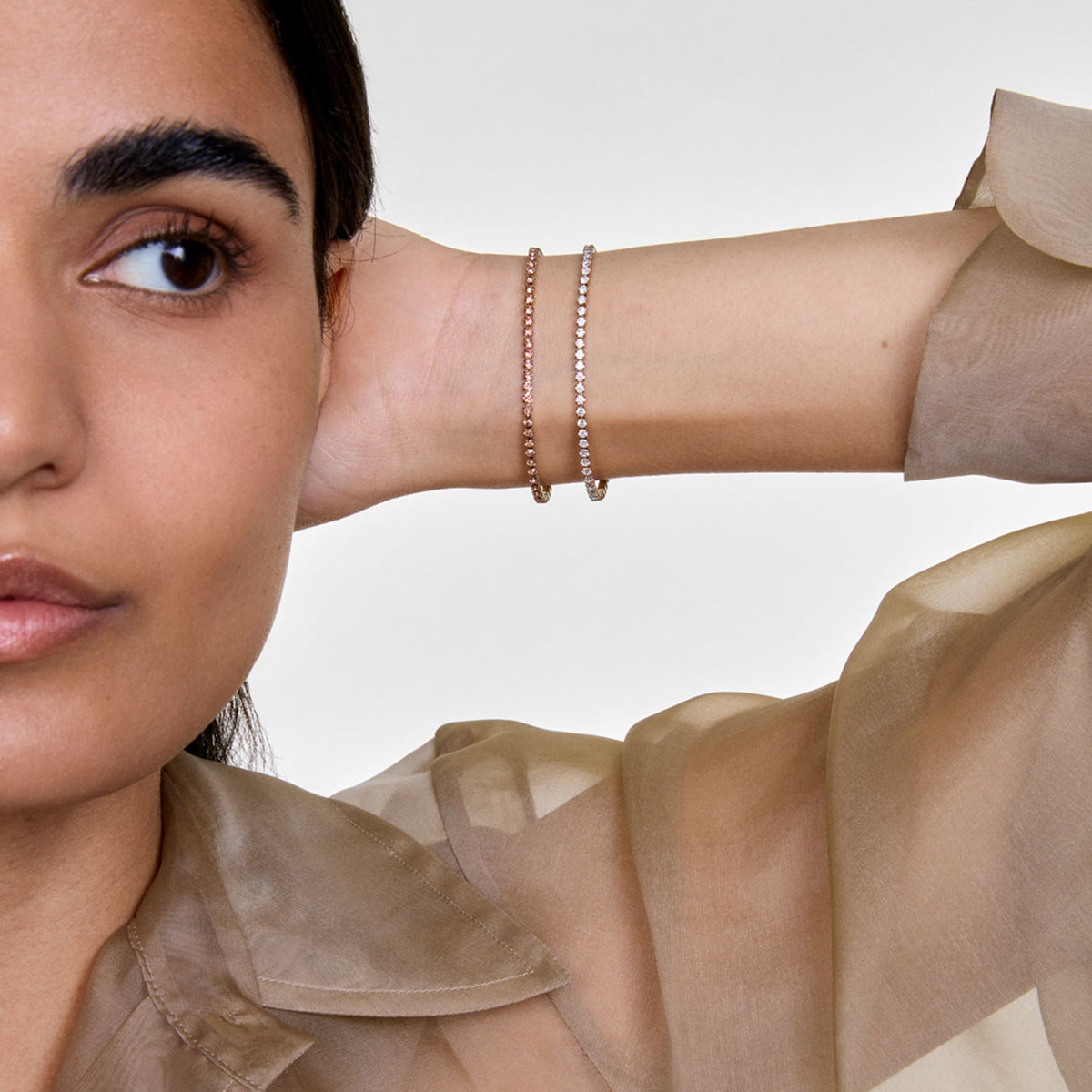 A woman wearing the Classic Tennis Bracelet Apricot Silver on her wrist, showcasing its slim silver band adorned with apricot-coloured crystals, styled with a sheer neutral blouse for an elegant look.