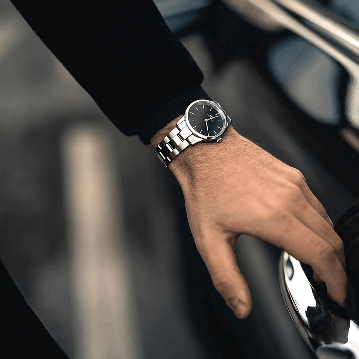 Daniel Wellington Iconic Link Silver watch with black dial and stainless steel strap, worn on wrist while opening a car door.