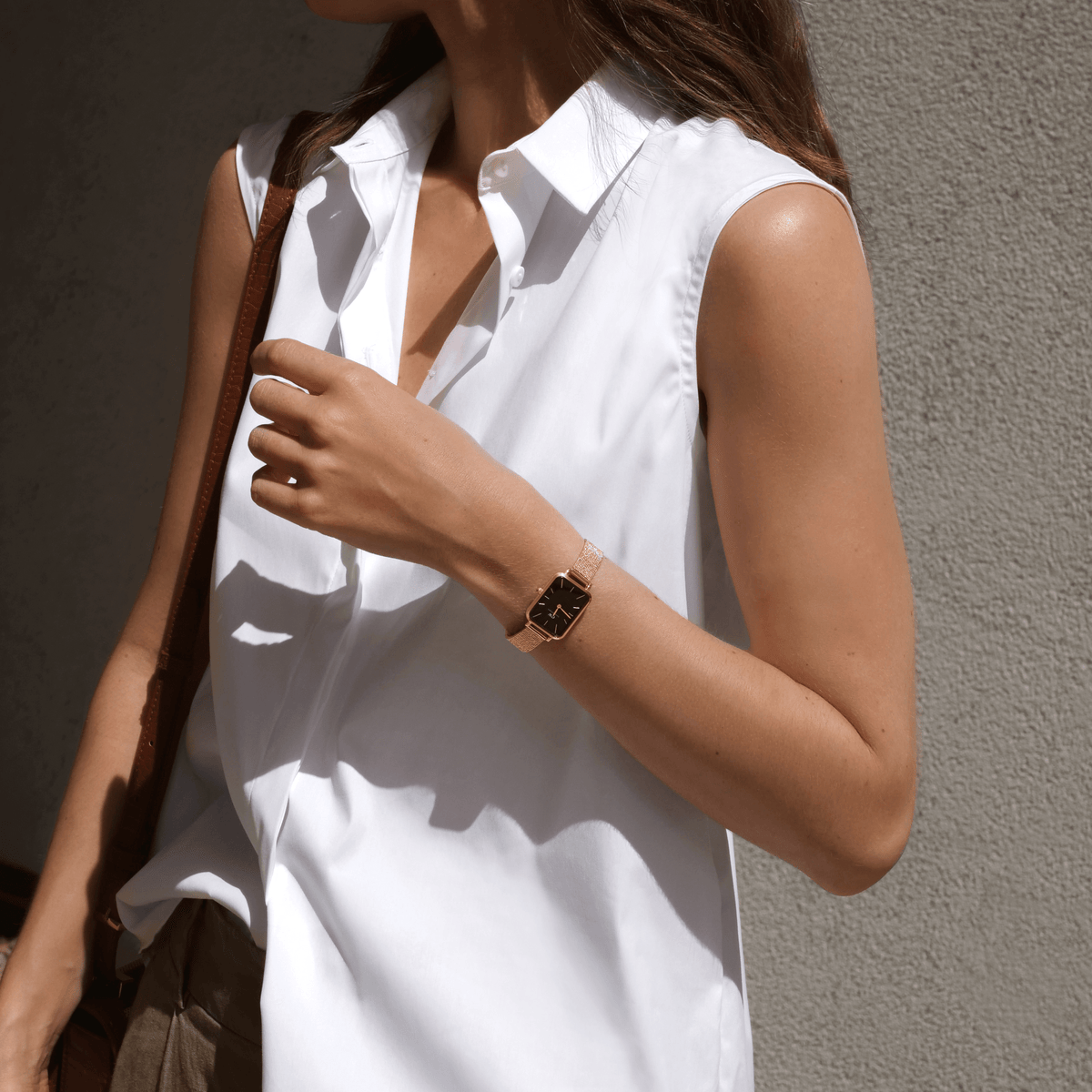 A woman wearing the Quadro Pressed Melrose Black Rose Gold watch with a rectangular black dial and rose gold mesh strap, styled with a sleeveless white shirt in natural sunlight.
