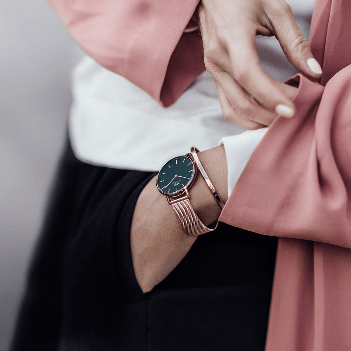 Petite Melrose watch with black dial and rose gold mesh strap styled on wrist, paired with a pink blazer and bracelet.