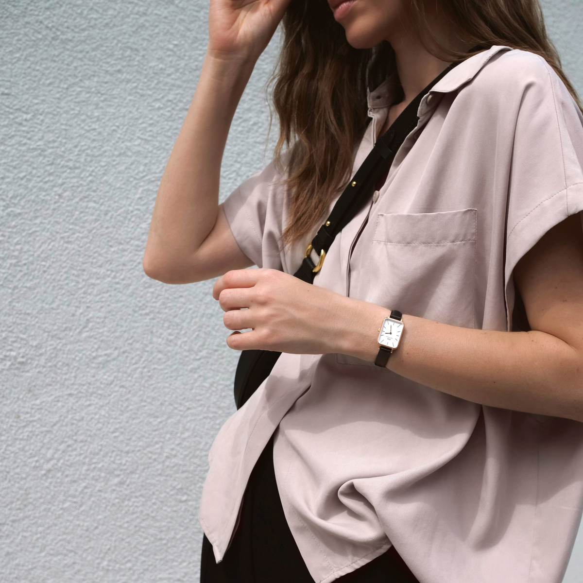 A woman wearing the Daniel Wellington Quadro Pressed Sheffield watch, featuring a rectangular eggshell white dial, rose gold case, and black leather strap, styled with a relaxed short-sleeve shirt and crossbody bag.