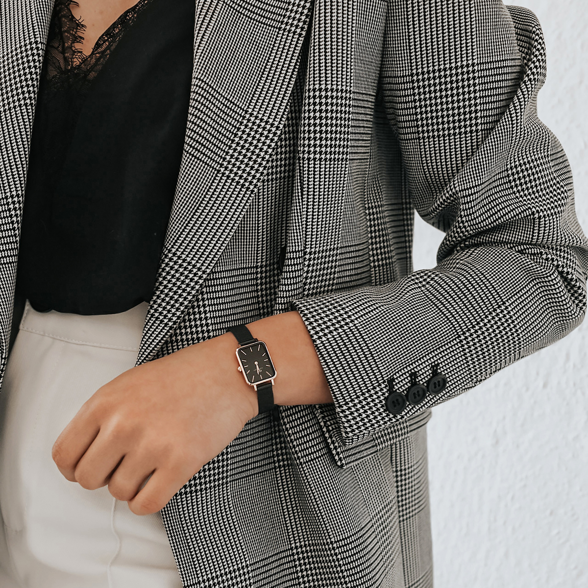 A woman wearing the Quadro Pressed Ashfield rectangular watch with a black dial, rose gold case, and black mesh strap, styled with a checked blazer and black lace top for a sophisticated, modern look.
