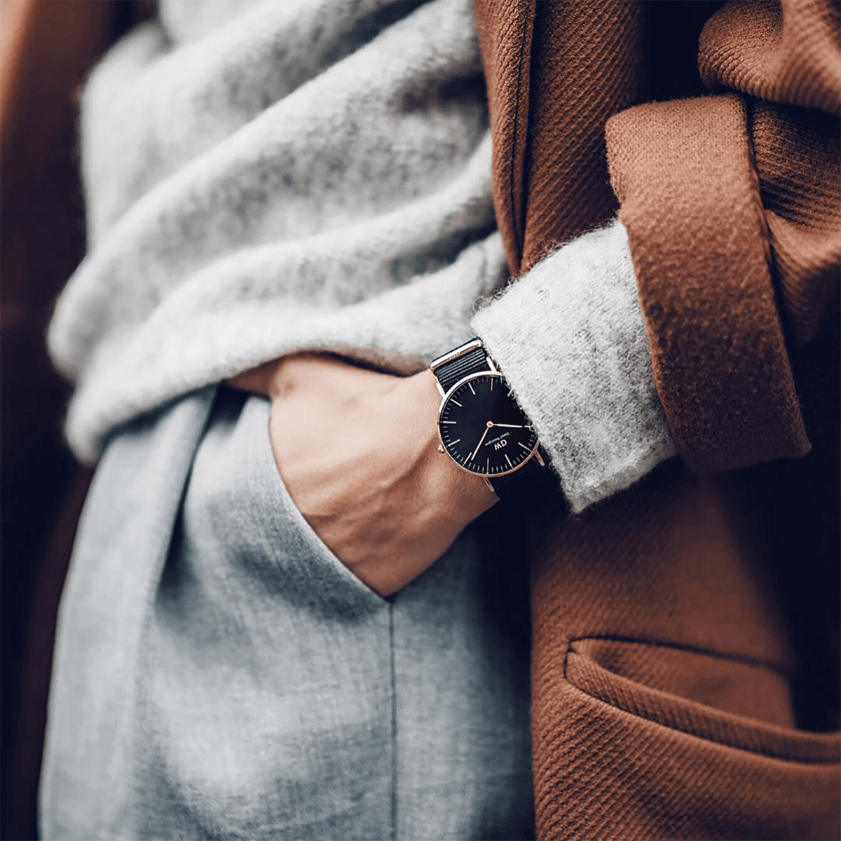 A person wearing the Classic Cornwall Black Rose Gold watch with a black dial and black NATO strap, styled with a grey jumper and brown coat for a smart casual look.