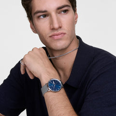 Man wearing the Daniel Wellington Classic Day Display Sterling Arctic watch, featuring a blue dial with day and date display and a polished silver mesh strap, styled with a dark collared shirt for a refined, modern look.