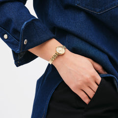A woman wears the Elan Lumine Unitone Gold watch with a gold 3-link bracelet and crystal-accented dial, styled with a dark denim shirt and black trousers.