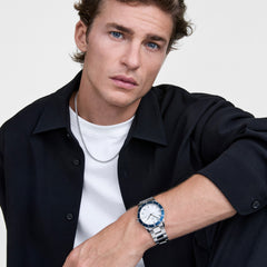 A man wearing the Daniel Wellington Iconic Paradigma Link Blue Bezel watch, featuring a polished stainless steel bracelet and a bold blue bezel, styled with a white t-shirt and black shirt for a modern, sophisticated look.