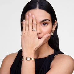 A woman wearing a sleeveless black top showcases a gold and silver square-faced watch on her wrist, paired with small gold hoop earrings and a pavé crystal ear cuff.