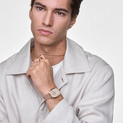 A man wearing the Daniel Wellington Marlon Wire Linen Rose Gold watch, featuring a cushion-shaped rose gold case, linen-textured dial, and matching wire mesh strap, styled with a light jacket and gold jewellery for a refined, modern look.