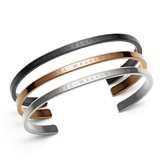 Set of three Daniel Wellington cuff bracelets in black, rose gold, and silver stainless steel, each featuring a minimalist open design with the brand name elegantly engraved along the band.