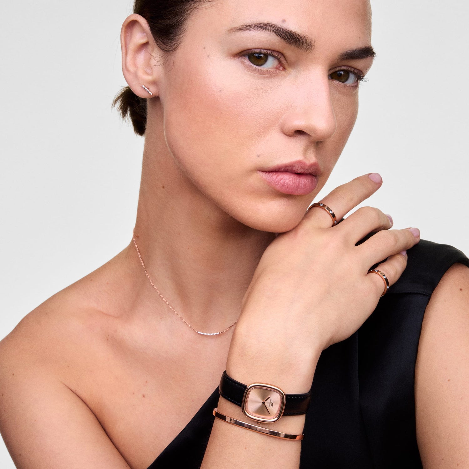 A woman wearing the Mirelle Eternity Necklace Rose Gold, featuring a fine rose gold chain and a curved bar set with white cubic zirconia crystals, styled with matching rose gold jewellery and a black watch for an elegant, minimalist look.
