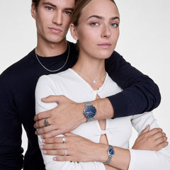 A couple wearing the Classic Day & Ophelia Mini Arctic Silver Set by Daniel Wellington, featuring a men’s blue dial mesh strap watch and a women’s petite blue dial silver bracelet watch, styled with coordinated jewellery for a modern, elegant look.