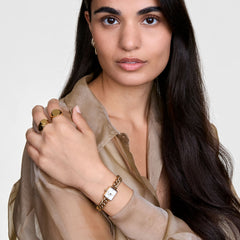 Woman wearing Jolie Chain Gold watch with chunky gold bracelet and white square dial, styled with gold rings and sheer blouse.