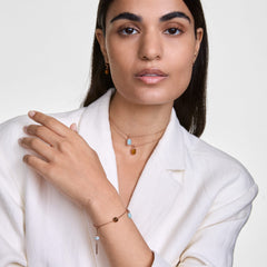 Woman wearing a white blazer styled with a rose gold charm extension chain on her bracelet and layered necklace, both featuring delicate charms in soft blue and amber tones for a modern, elegant look.