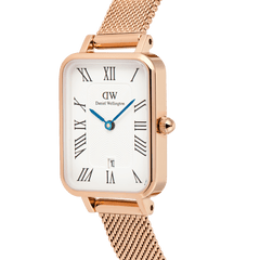 Close-up of the Quadro Melrose women’s watch with a rectangular rose gold case, white dial featuring Roman numerals, blue hands, guilloché detailing, and a rose gold mesh strap. The date window is positioned at 6 o’clock for added practicality.
