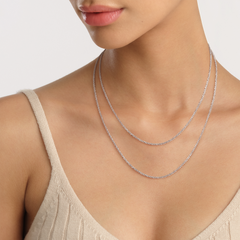 A woman wears the Twisted Chain Necklace S, a delicate silver chain with a twisted design, layered elegantly at the collarbone over a neutral knit top.