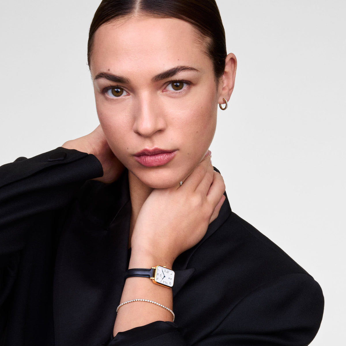 A woman wearing the Quadro Roman Numerals Date Sheffield Gold watch, featuring a rectangular gold case, white dial with Roman numerals, blue hands, and a black leather strap, styled with a black blazer and a delicate bracelet.