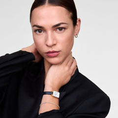 A woman wearing the Quadro Roman Numerals Date Sheffield Gold watch, featuring a rectangular gold case, white dial with Roman numerals, blue hands, and a black leather strap, styled with a black blazer and a delicate bracelet.