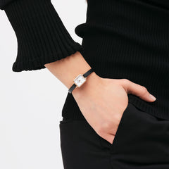 A woman wears the Quadro Mini Sheffield Rose Gold watch with a rectangular rose gold case, white dial, and slim black leather strap, styled with a black ribbed top and trousers.