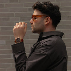A man wearing the Iconic Chronograph St Mawes Amber RG watch with a rose gold case, amber dial, and brown leather strap, styled with a dark patterned shirt against a brick wall backdrop.